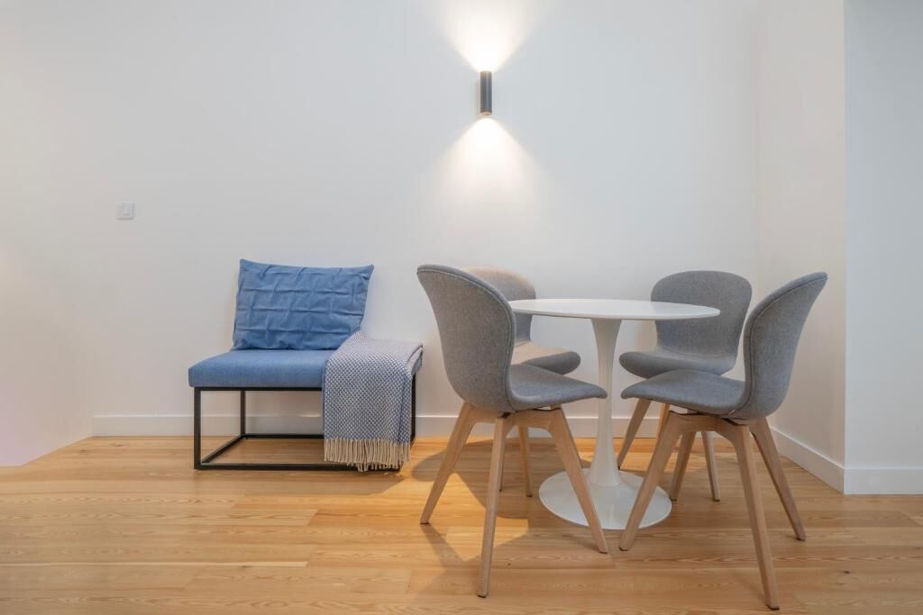 Une pièce avec une table ronde et quatre chaises, un canapé bleu avec un coussin et un luminaire fixé au mur.