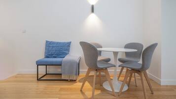 Una habitación pequeña y moderna con una mesa redonda y dos sillas grises, un sofá azul con un cojín a juego, y una lámpara fijada a la pared.