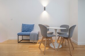 A small, modern room with a round table and two grey chairs, a blue sofa with a matching cushion, and a wall-mounted light fixture.