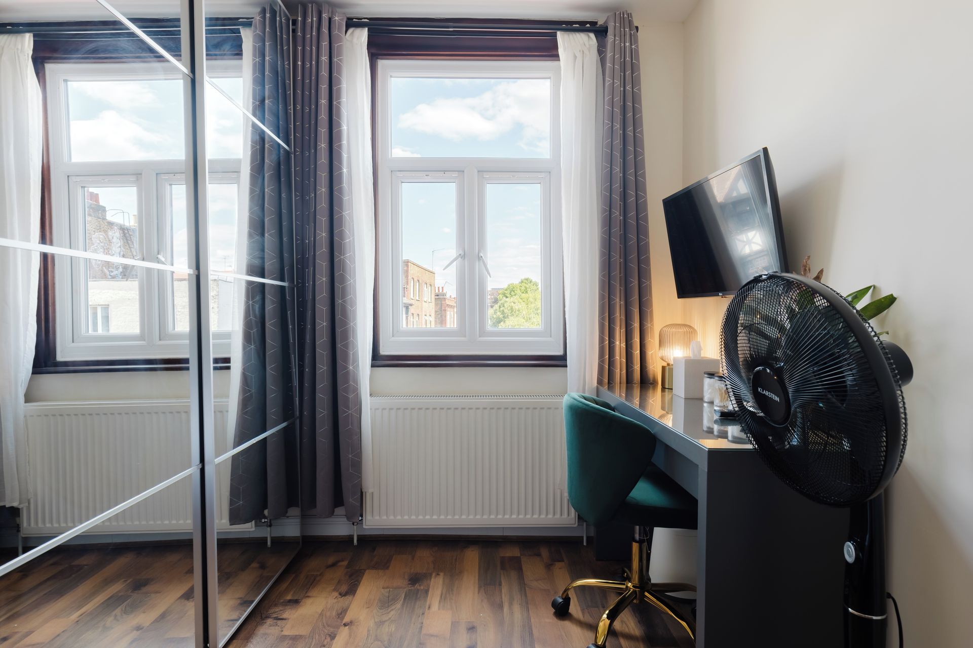 A room with a desk, a chair, a television, a radiator, and a window with curtains.