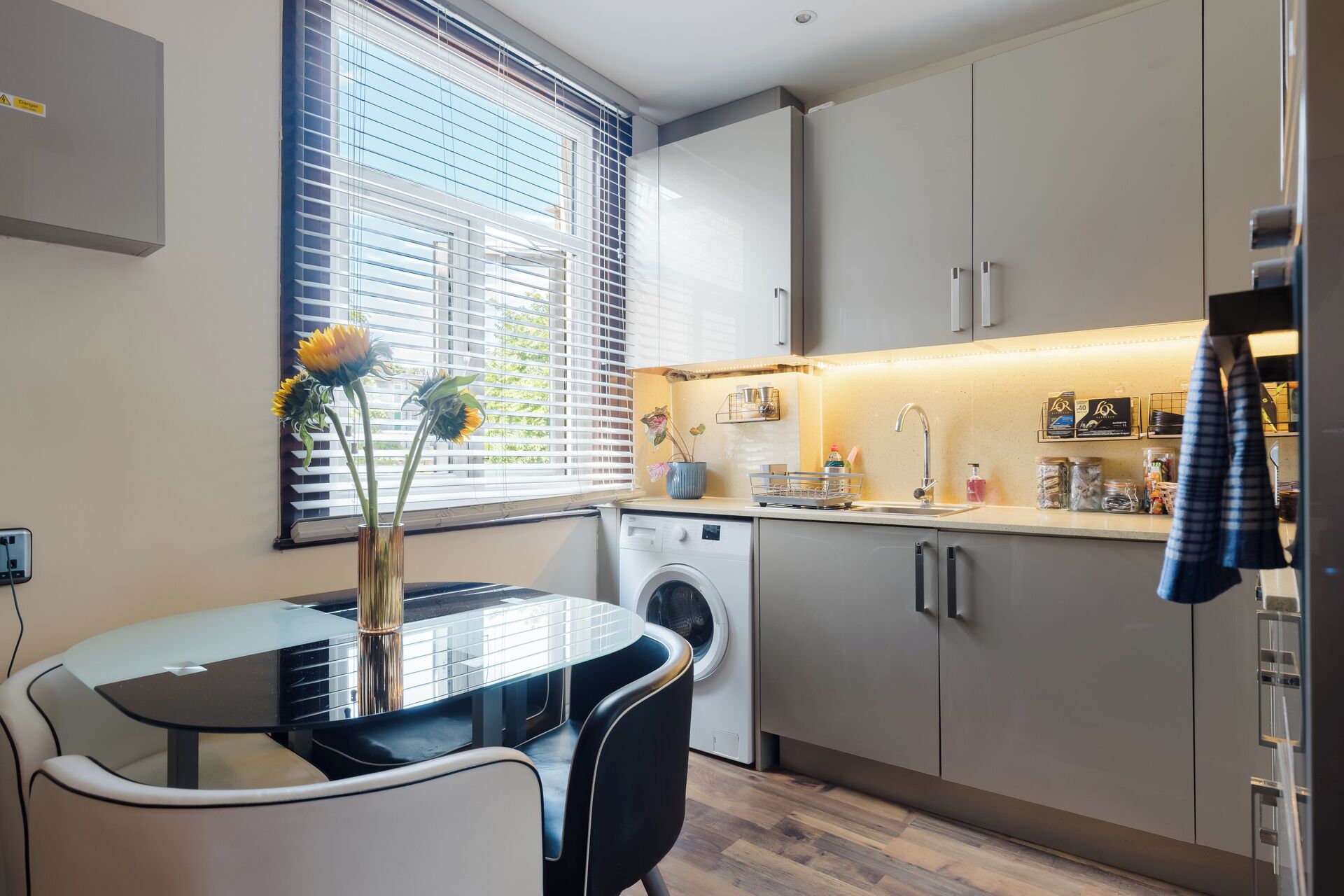 A compact kitchen with a washing machine, a dining table with chairs, and a window with blinds.