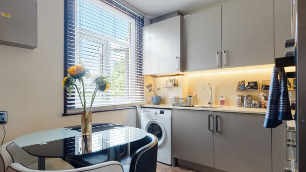 Een praktische keuken met een wasmachine, een eettafel met stoelen en een raam met jaloezieën.