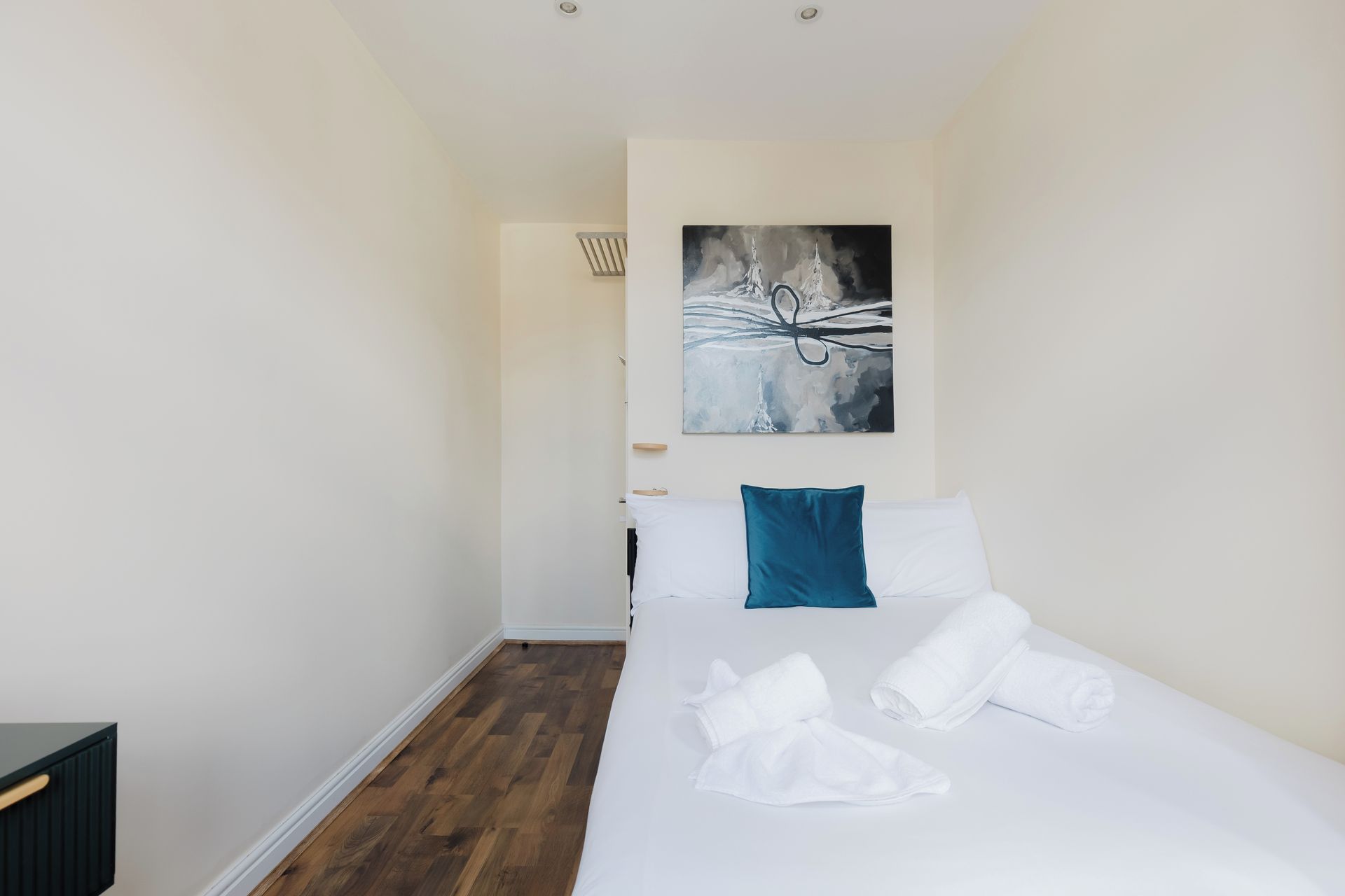 A hotel room with a bed, white linens, a blue pillow, and a painting on the wall.