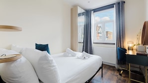 Een hotelkamer met een bed, een bureau, een lamp en een raam met uitzicht op gebouwen.