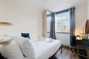 Ein Hotelzimmer mit Bett, Schreibtisch, Lampe und einem Fenster mit Blick auf Gebäude.
