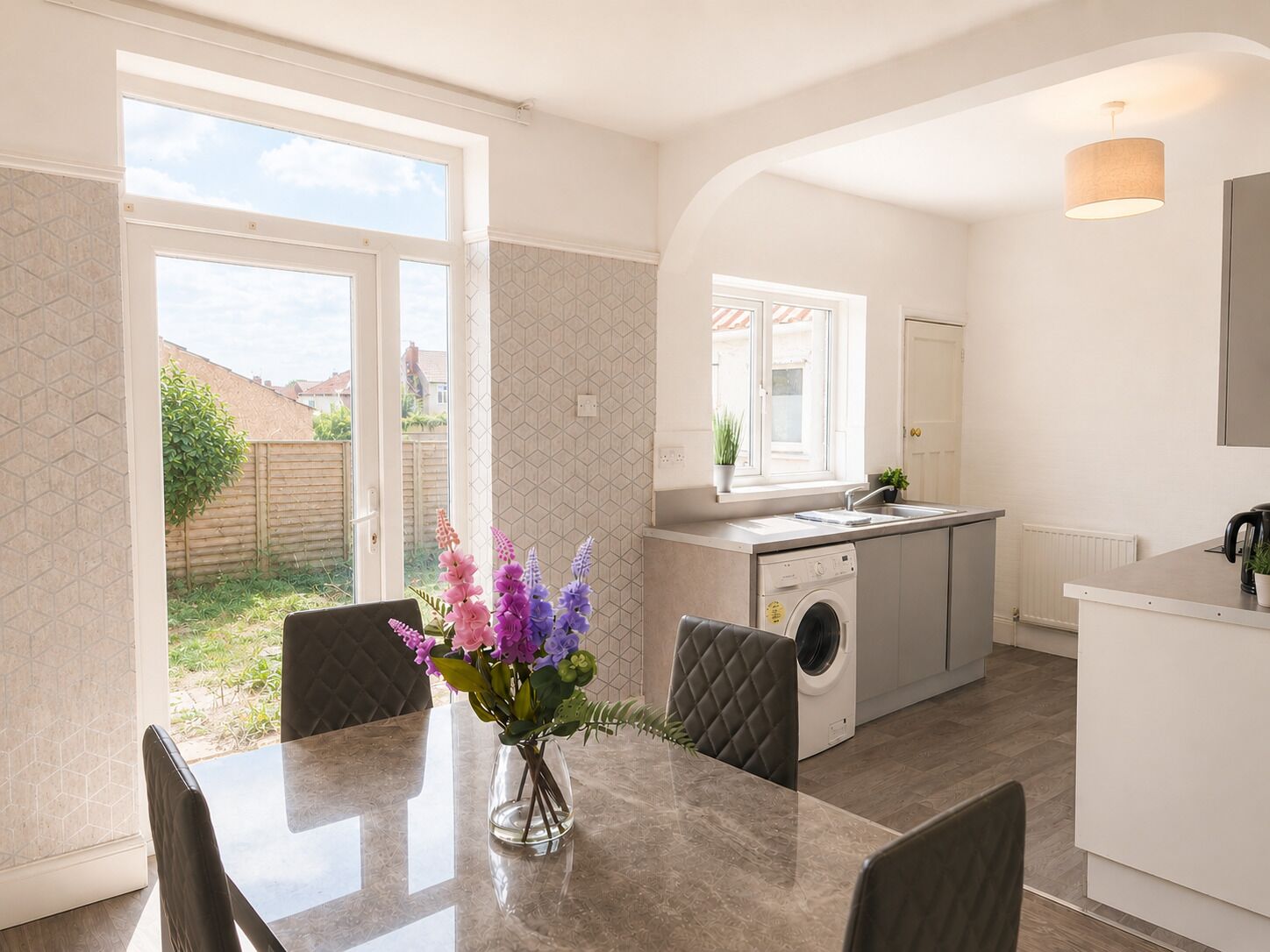 Une cuisine moderne équipée d’un lave-linge intégré, un coin repas avec une table et des chaises, et une vue sur le jardin à travers une porte vitrée.