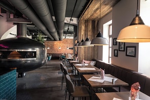 Interior de restaurante com parede de tijolos, teto de madeira e uma grande churrasqueira. Mesas e cadeiras dispostas para refeições.