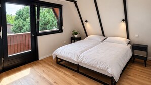 A bedroom with a large bed, a wooden floor, a window with a view of greenery, and a door leading outside.
