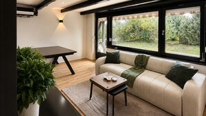 A modern living room with a sofa, a coffee table, and a dining table. There are large windows providing a view of greenery outside.