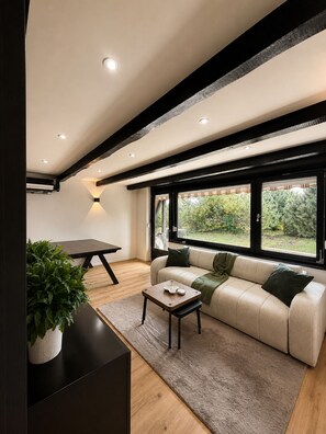Ein modernes Wohnzimmer mit einer Couch, einem Couchtisch und einem Esstisch. Große Fenster bieten Blick auf die grüne Umgebung.