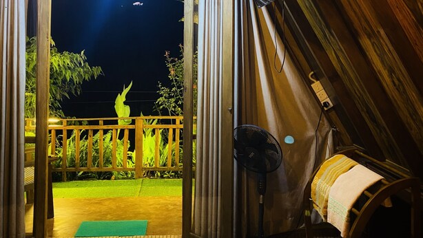 Ein Dachzimmer mit Holzbalkendecke, einem Bett mit gemustertem Überwurf, einem Standventilator und Blick auf einen Garten bei Nacht.