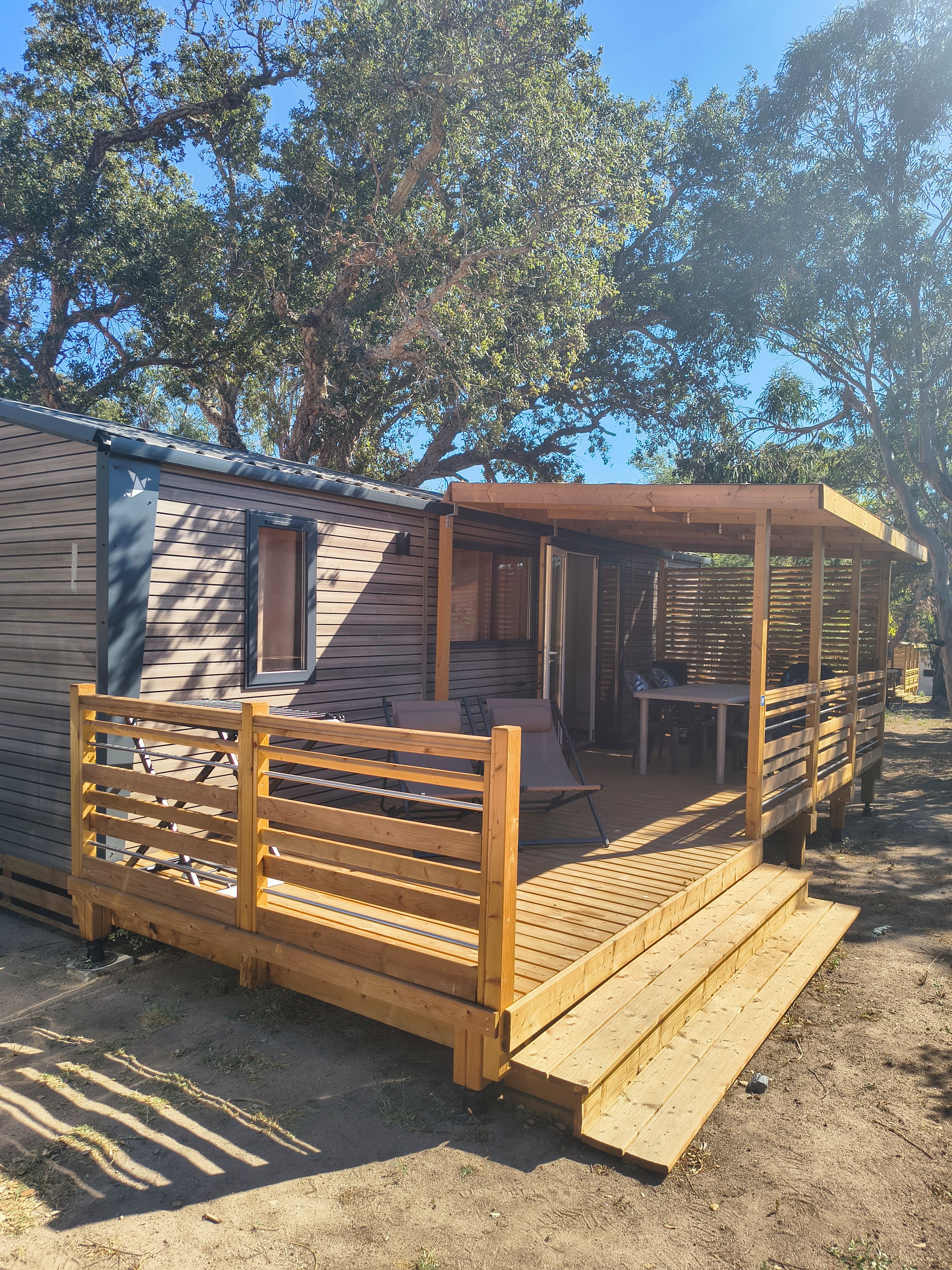 Eine Holzterrasse mit einer Bank und einem Tisch, ein überdachter Patio-Bereich und ein Wohnwagen mit einem Fenster.