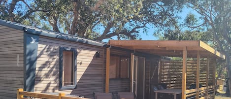 Eine Holzterrasse mit einer Bank und einem Tisch, ein überdachter Patio-Bereich und ein Wohnwagen mit einem Fenster.