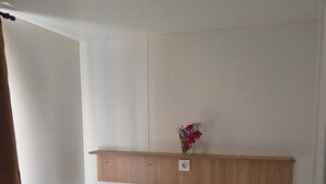 Un lit simple avec une étagère en bois au-dessus, un petit vase avec des fleurs et une fenêtre ornée d’un rideau marron.