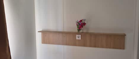 Una cama individual con un estante de madera encima, un jarrón con flores y una ventana con cortina marrón.