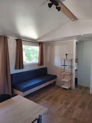 A room with a wooden floor, a dark blue bench, a white shelf unit, and a window with brown curtains.