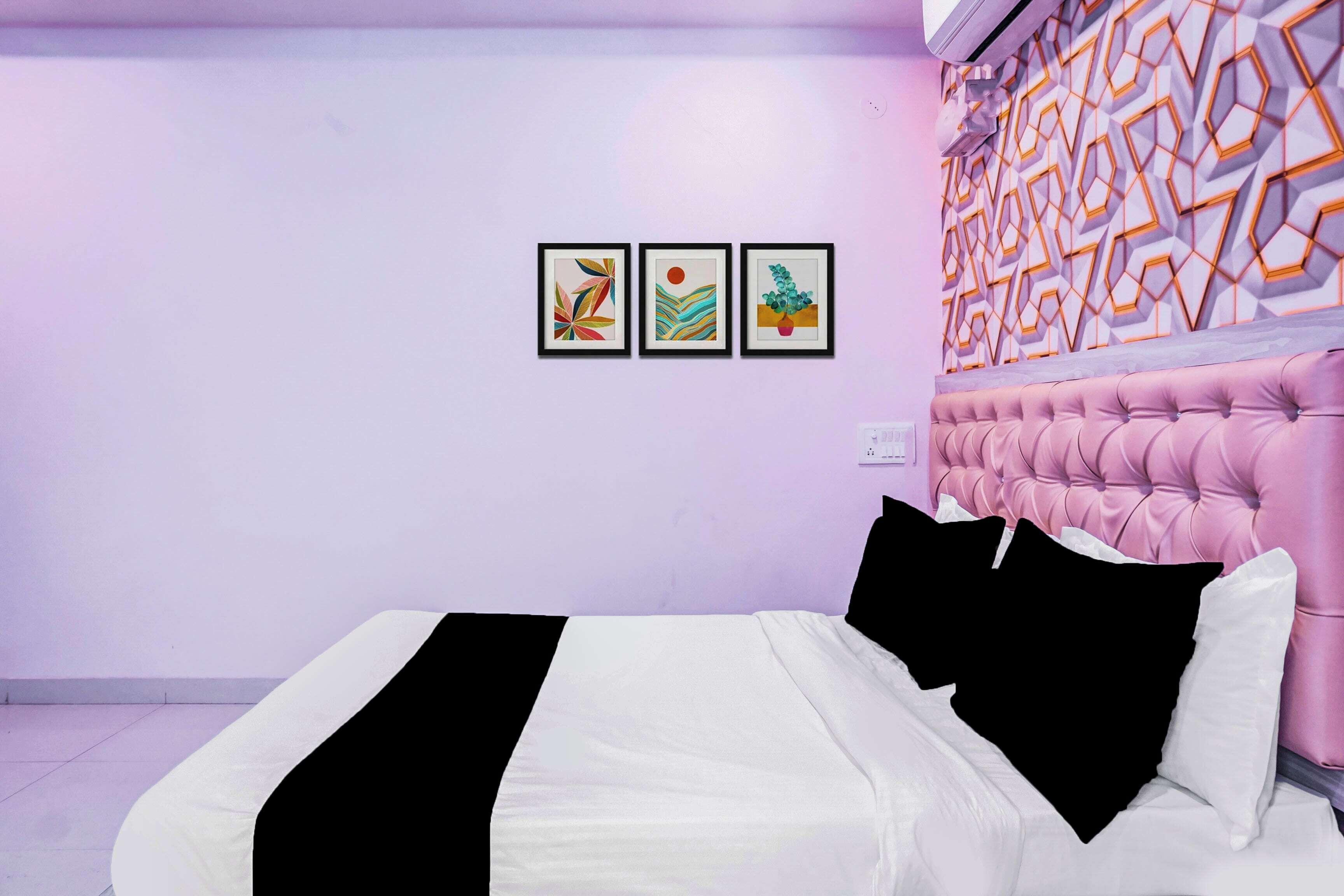 Une chambre à coucher avec une tête de lit rose à motifs capitonnés, un lit avec une literie blanche et noire, et trois œuvres d’art encadrées accrochées au mur.