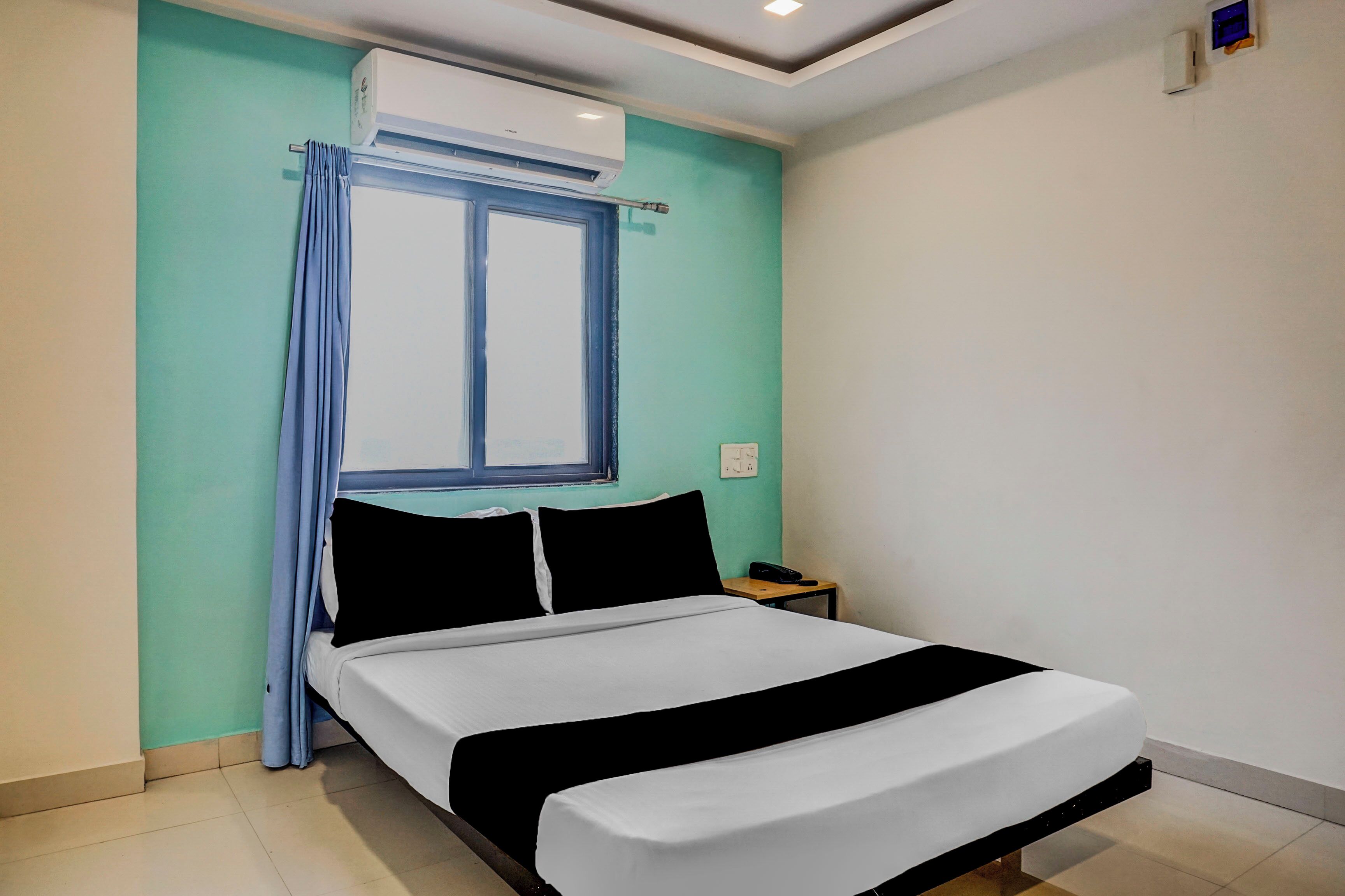 A hotel room with a bed, a window with curtains, an air conditioning unit, a bedside table with a phone, and a wall socket.