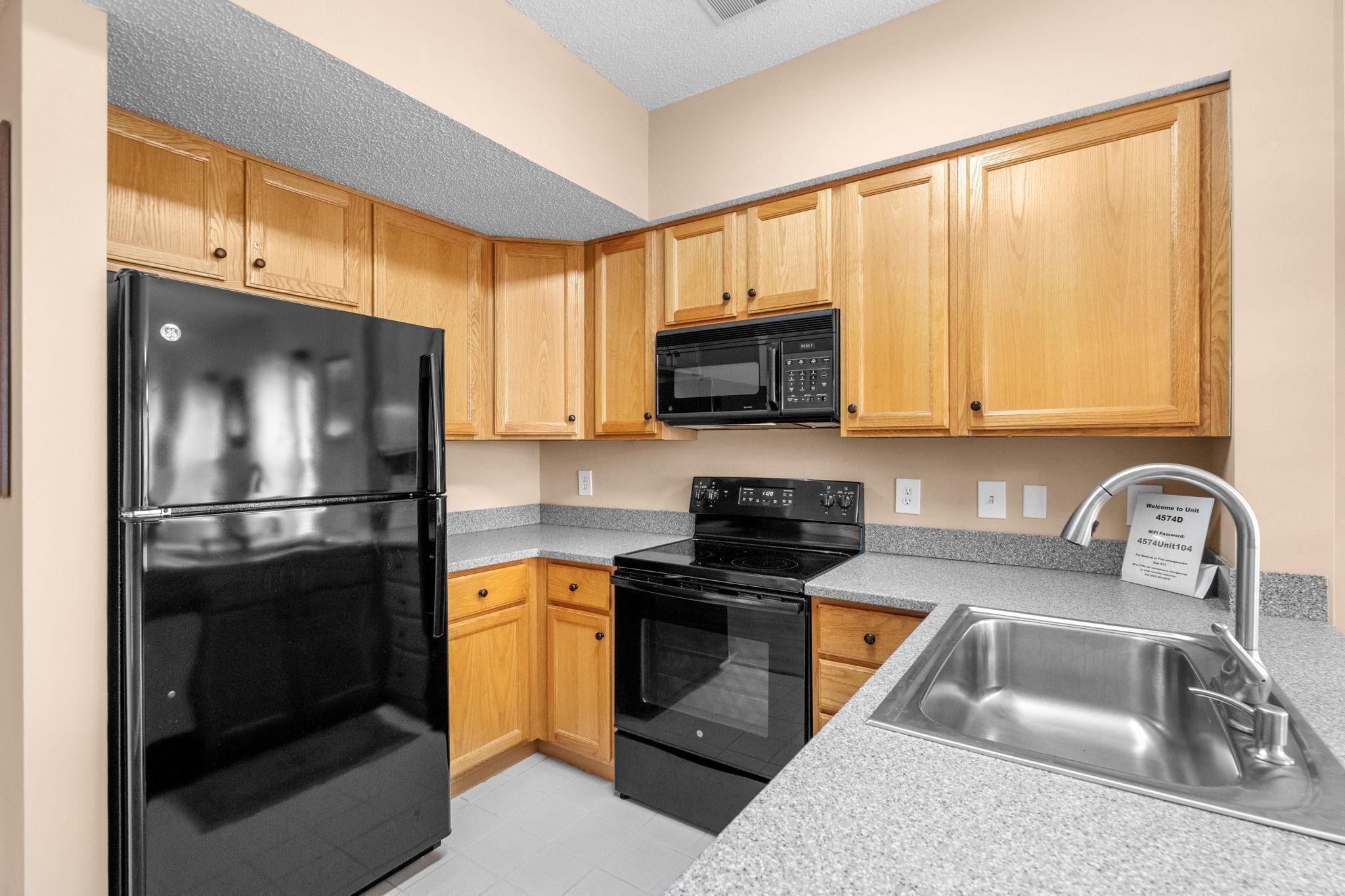 Una cocina con armarios de madera, un refrigerador negro, un microondas, un horno y un lavamanos de acero inoxidable.
