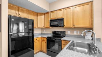Una cocina con armarios de madera, un refrigerador negro, un microondas, un horno y un lavamanos de acero inoxidable.