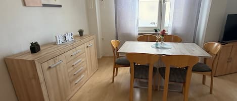 Un coin repas avec une table et des chaises en bois, un meuble avec des tiroirs, un tableau accroché au mur et une fenêtre avec des rideaux.