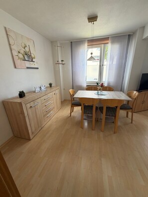 Un comedor con una mesa y sillas de madera, un armario con cajones, un cuadro en la pared y una ventana con cortinas.