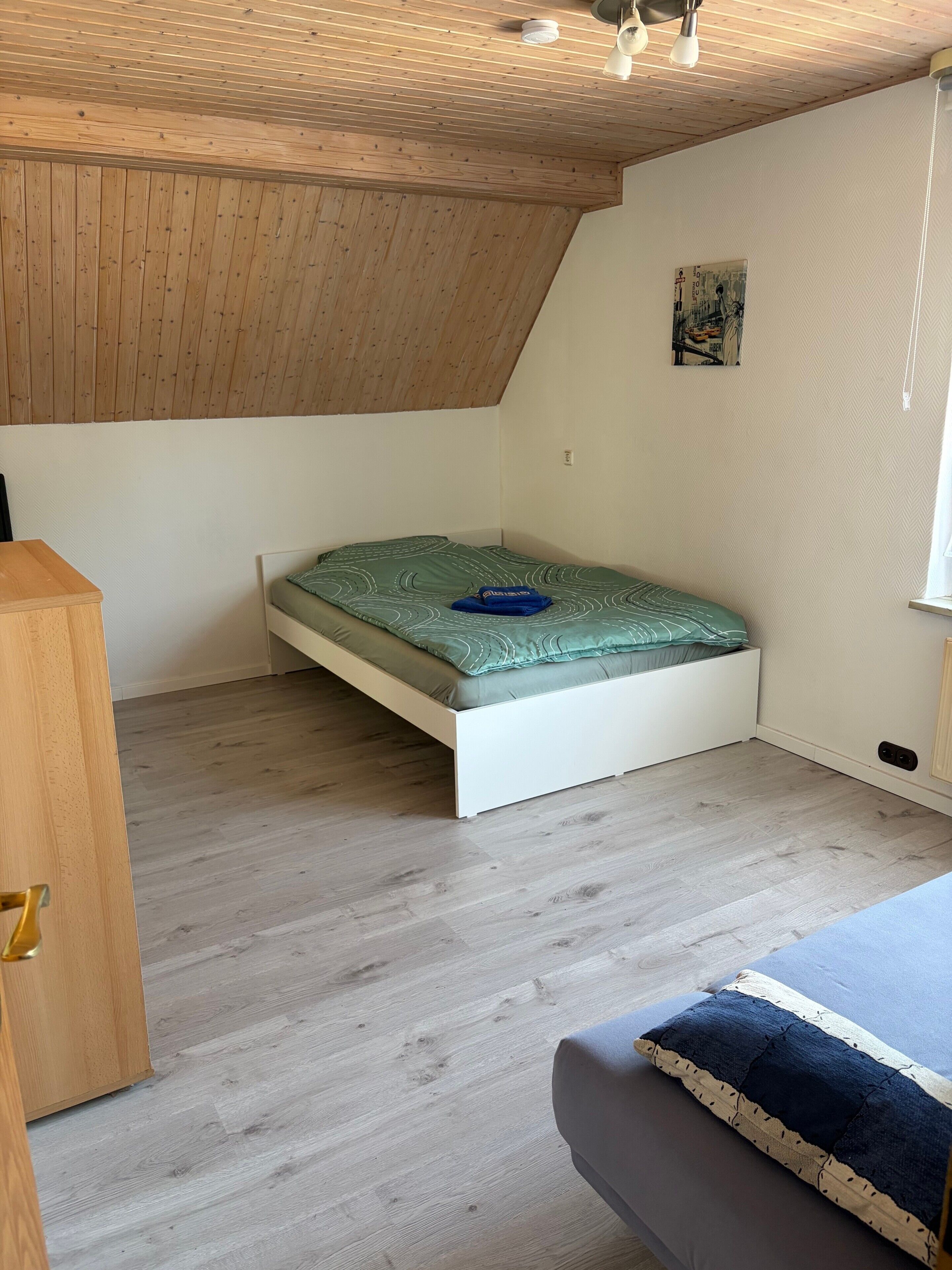 A room with a sloped ceiling, a bed with a green mattress, a wooden nightstand, and a window.