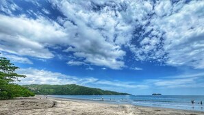 Una playa con cielo despejado y mar tranquilo, rodeada de vegetación y colinas a lo lejos.