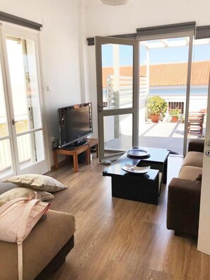 A living room with a sofa, a coffee table, a television, and a sliding glass door leading to an outdoor area with potted plants. - The Roof in Aegina Town (Aegina)