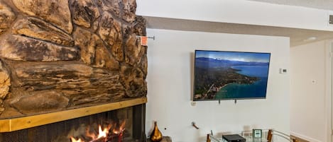 A stone fireplace with a visible fire, a mounted television on the wall, and a glass table with a metal frame.