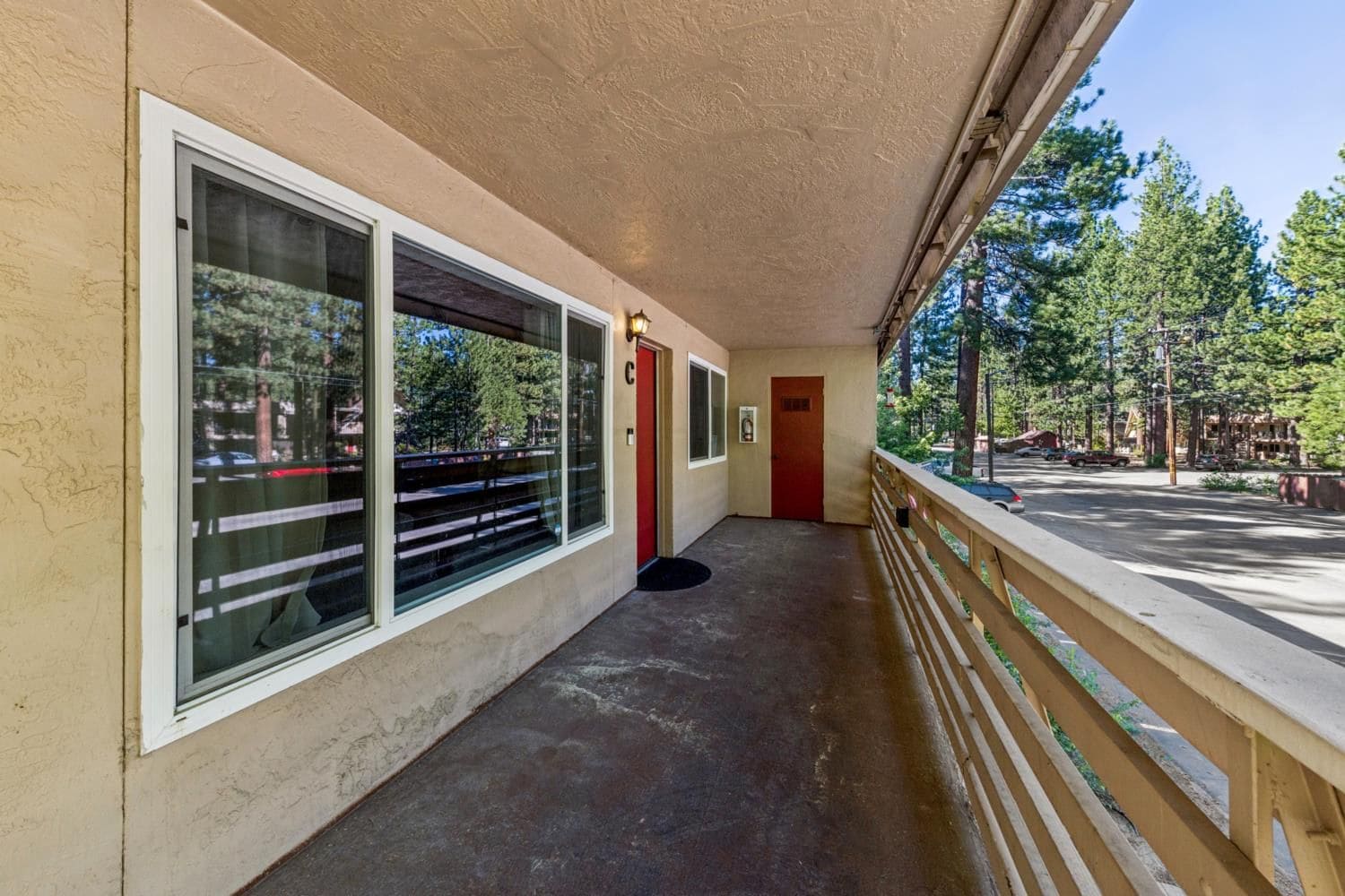 A covered corridor with large windows, a red door, and a wooden railing overlooking a parking area.