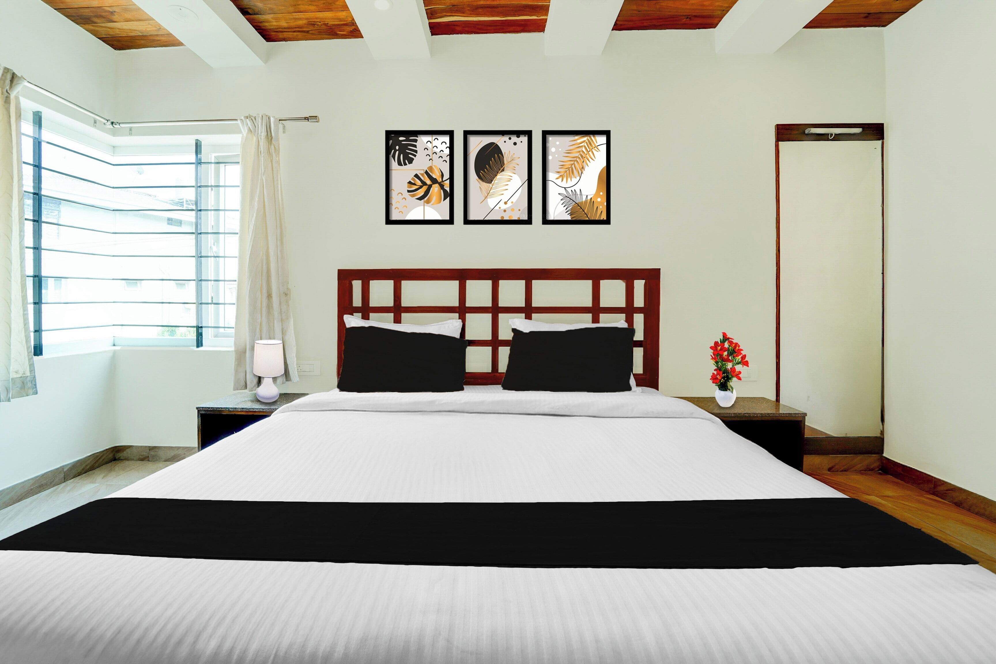 A hotel room with a large bed, a nightstand with a lamp, a window with curtains, and three framed artworks on the wall.