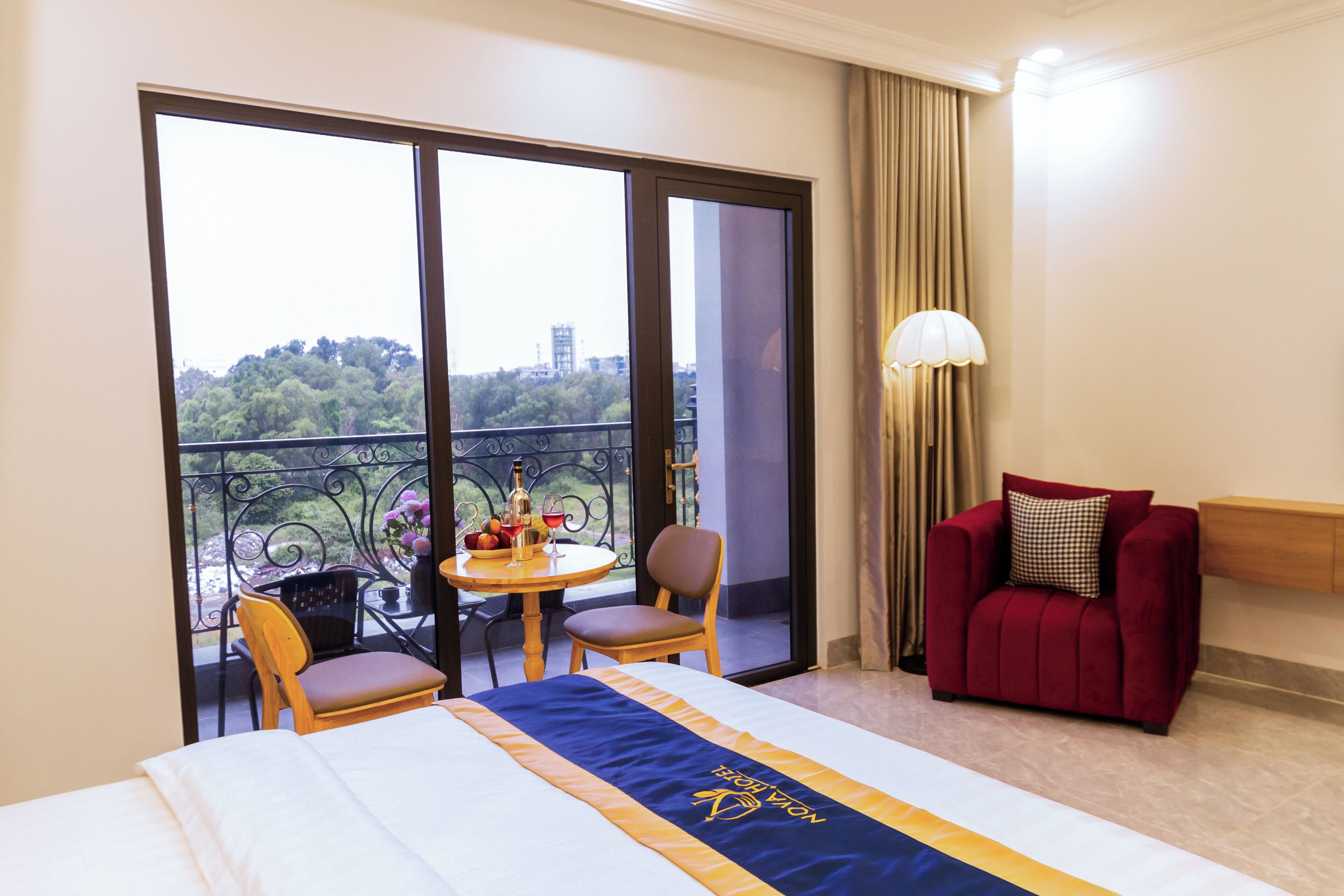 Habitación de hotel con cama, una mesa y sillas, un sillón rojo y vistas a un balcón con plantas y edificios.