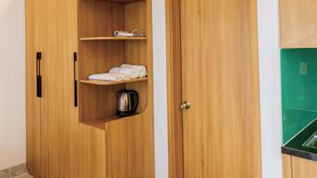 A room with a wooden wardrobe, a built-in shelf with towels, a kettle, and a pair of slippers on the floor.