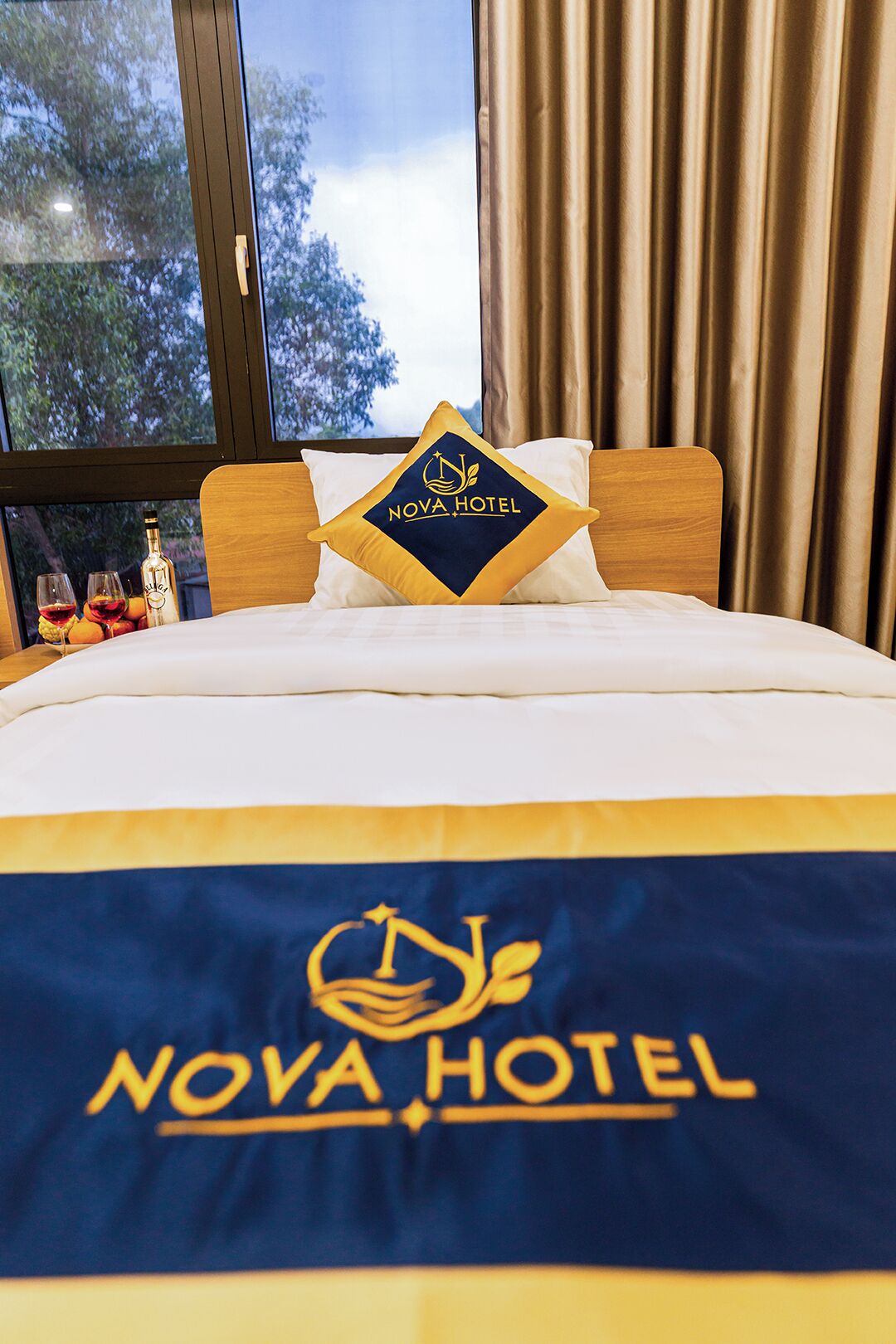 Una cama con edredón que presenta el logotipo del Hotel Nova, una ventana con cortinas y vistas a árboles fuera.