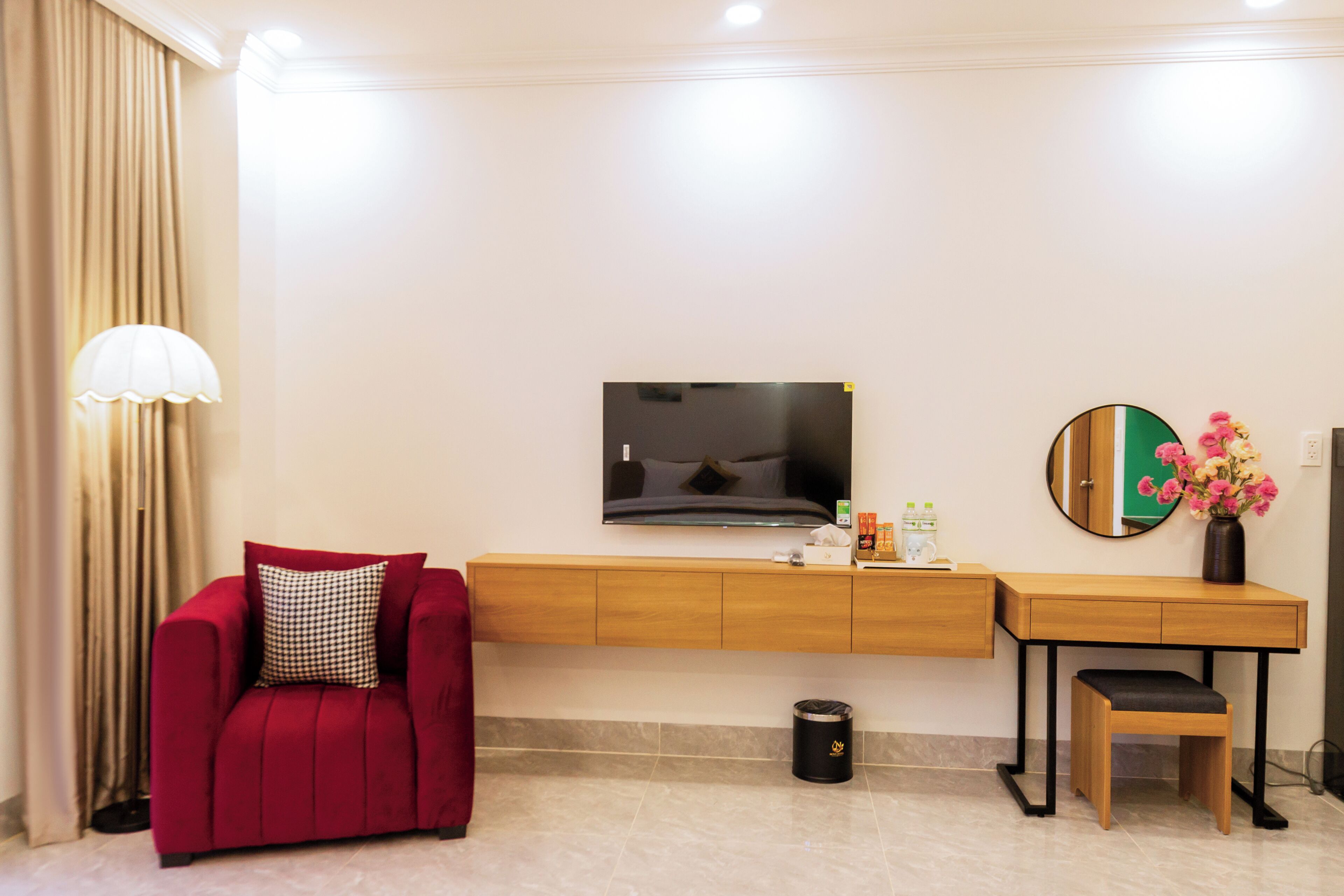 Une chambre d’hôtel avec un canapé rouge, une console en bois, une télévision, une petite poubelle et un vase avec des fleurs.