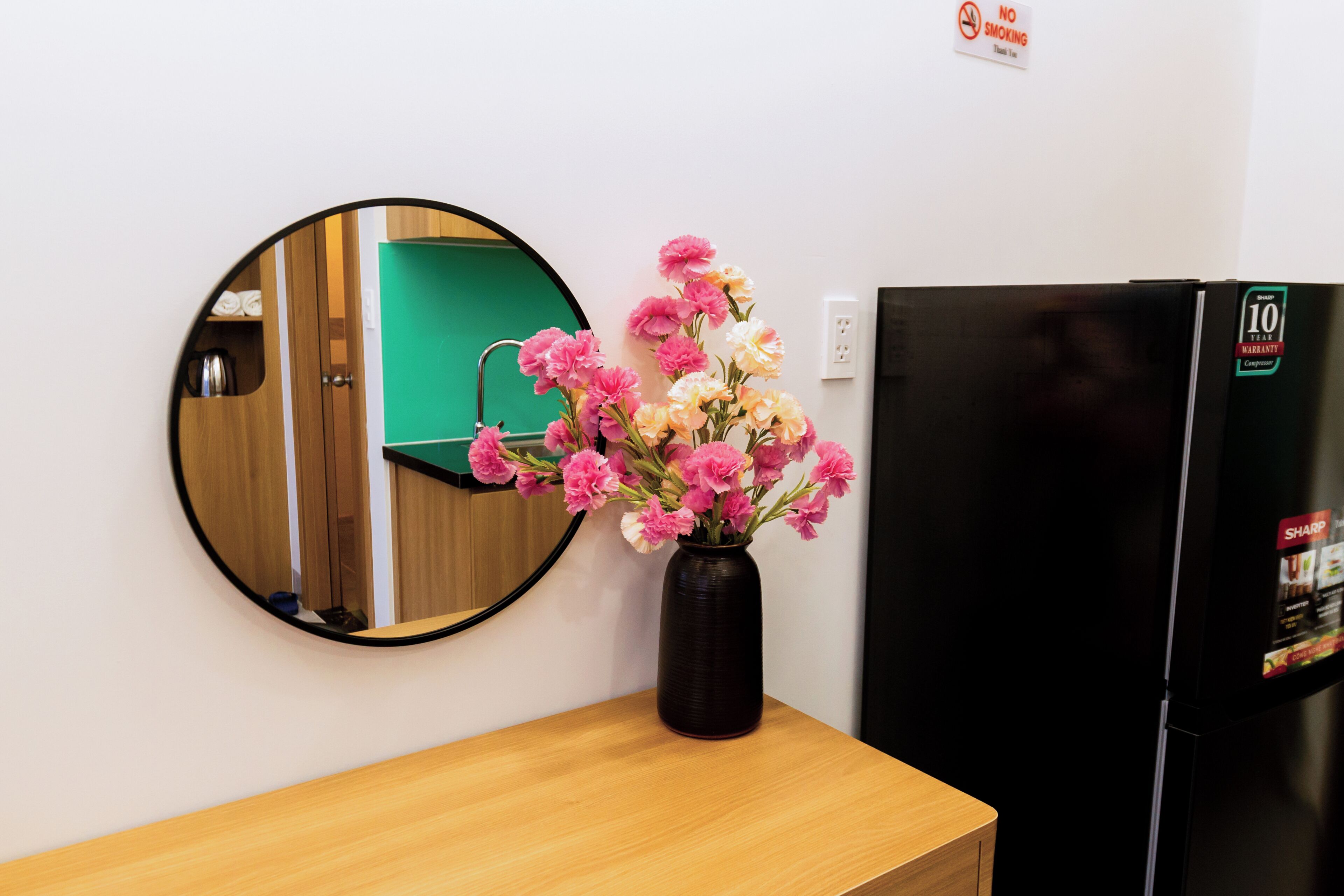 A room with a round mirror, a black refrigerator, and a wooden table with a vase of flowers.