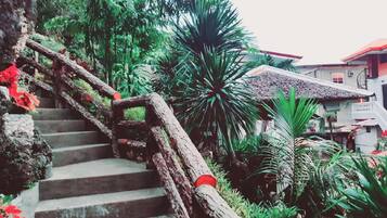 Una escalera con barandilla de madera rústica, rodeada de abundante vegetación y una casa con tejado rojo al fondo.