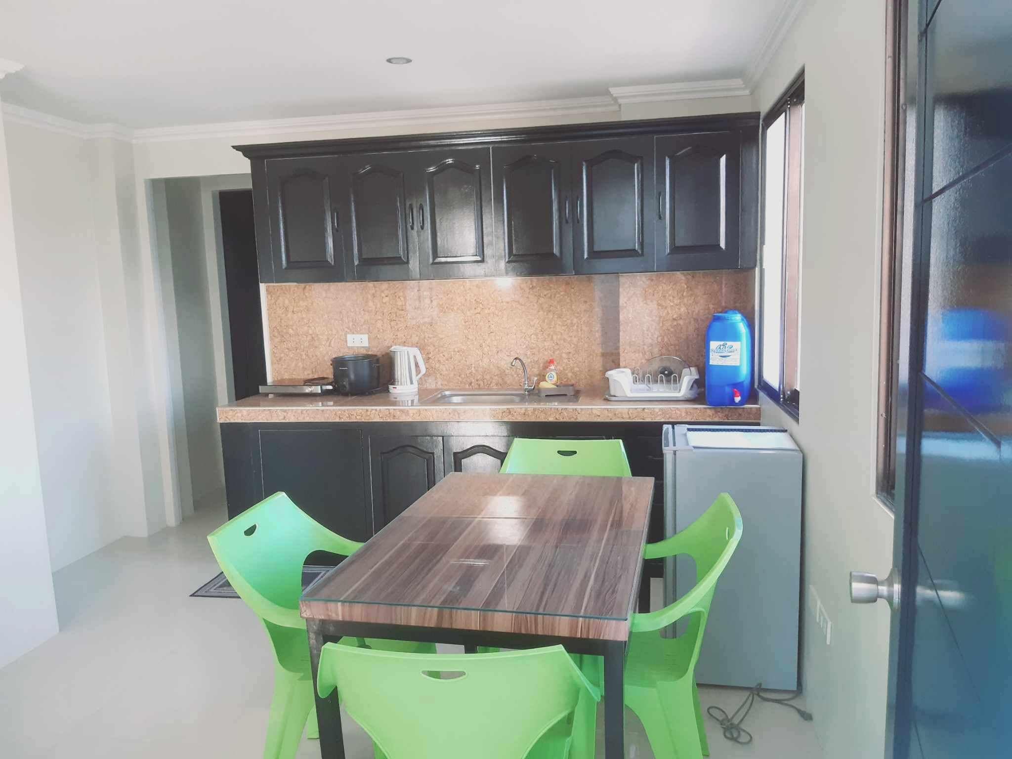 Una cocina compacta con armarios oscuros, una encimera, un lavabo, una cocina y una pequeña nevera, además de una mesa de comedor con sillas verdes.