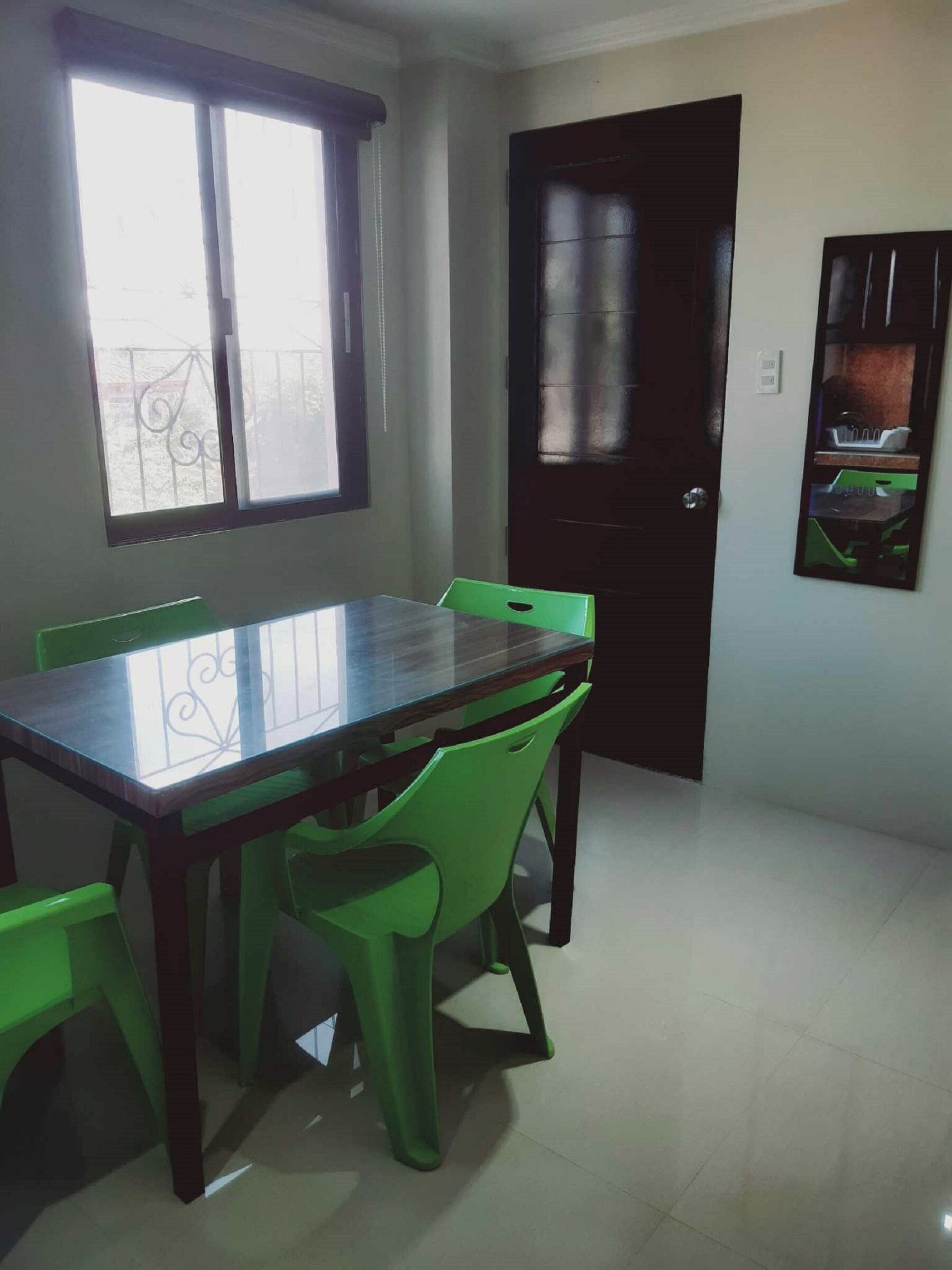 A dining area with a glass-top table and green chairs, a window with a metal railing, and a dark door.