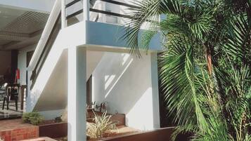 Un edificio de dos pisos con fachada blanca, un nivel inferior de ladrillo y un espacio al aire libre cubierto con escaleras que suben a un balcón.