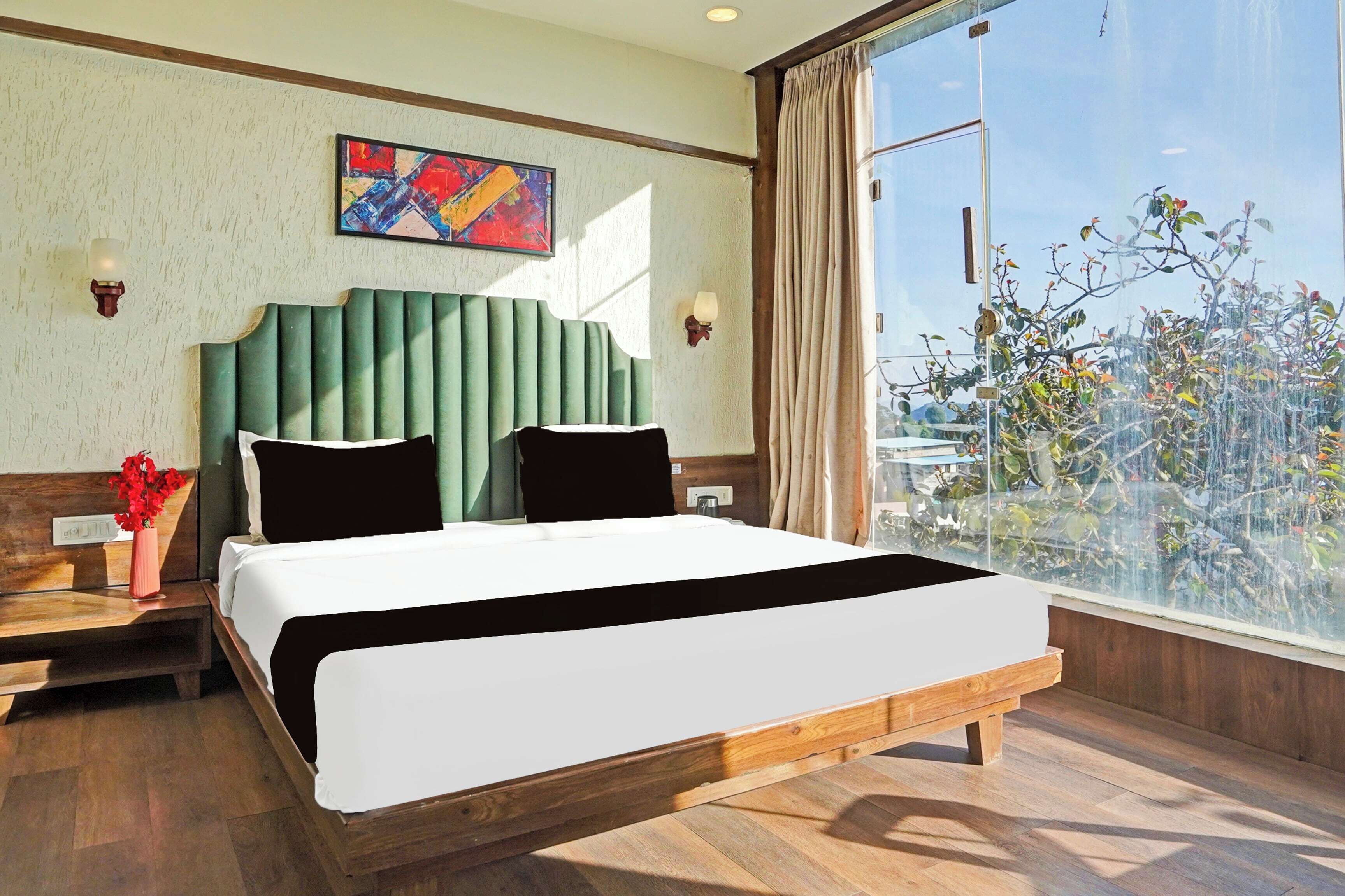 Habitación de hotel con una cama grande, una mesita de noche, un jarrón con flores, un cuadro en la pared y vistas a árboles a través de una ventana.