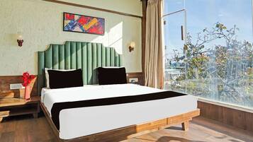 Habitación de hotel con una cama grande, una mesita de noche, un jarrón con flores, un cuadro en la pared y vistas a árboles a través de una ventana.