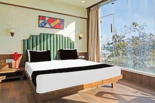 A hotel room with a large bed, a nightstand, a vase with flowers, a painting on the wall, and a view of trees through a window.