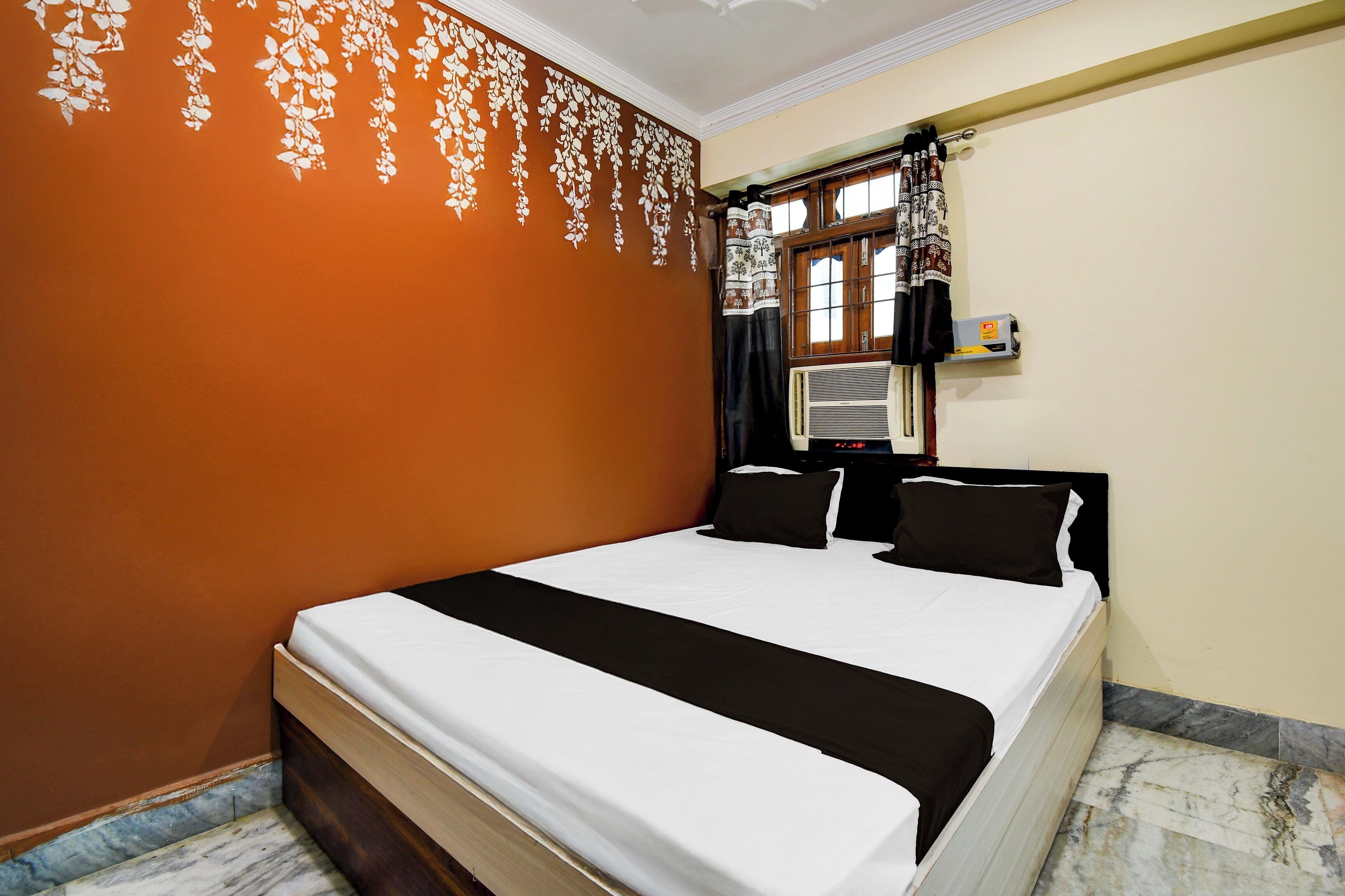 Una habitación de hotel con cama, ventana con cortinas, equipo de aire acondicionado y una pared con estampado floral.