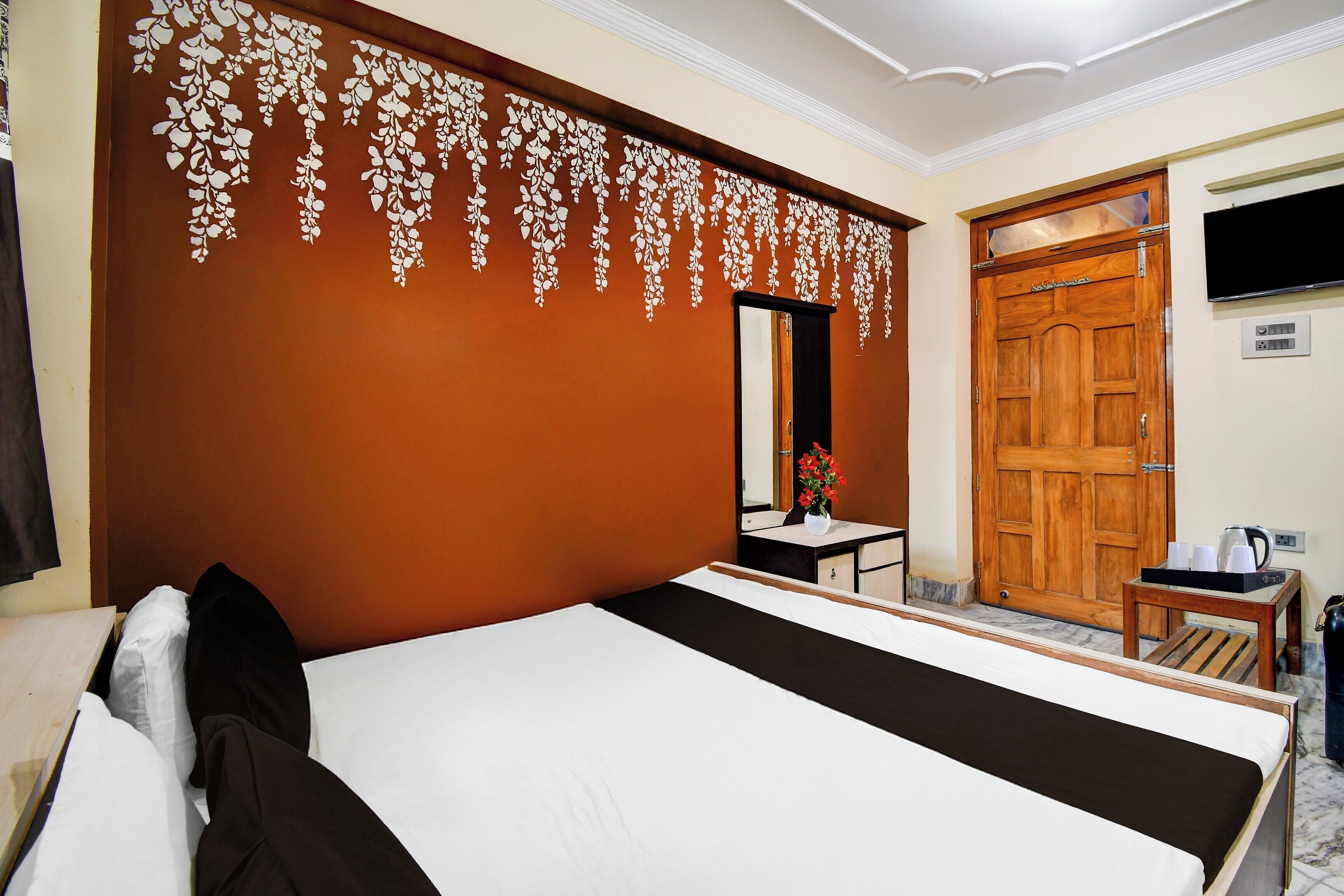 A hotel room with a bed, a small table with a vase of flowers, a wooden door, and a television mounted on the wall.