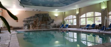 An indoor swimming pool with a stone wall, lounge chairs, and large windows.