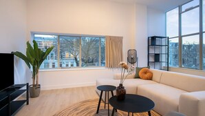 A modern living room with a sofa, a coffee table, a TV, and a large window offering a city view. - Modern 2 Bedroom Apartment in Rotterdam City Center With Free Parking (Rotterdam)