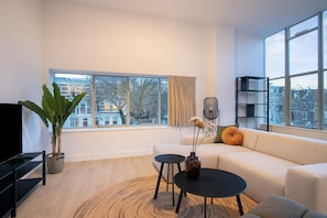 A modern living room with a sofa, a coffee table, a TV, and a large window offering a city view. - Modern 2 Bedroom Apartment in Rotterdam City Center With Free Parking (Rotterdam)