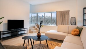 Ein modernes Wohnzimmer mit Flachbildfernseher, einer Couch, zwei kleinen Tischen und Blick auf eine Skyline durch große Fenster.